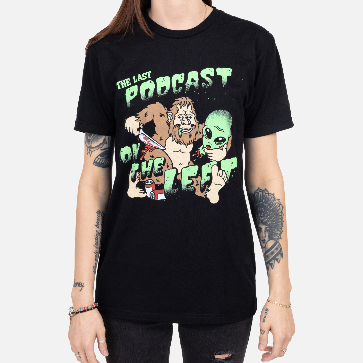 Black tee with graphic of bigfoot and alien head with text "THE LAST PODCAST ON THE LEFT" on female model