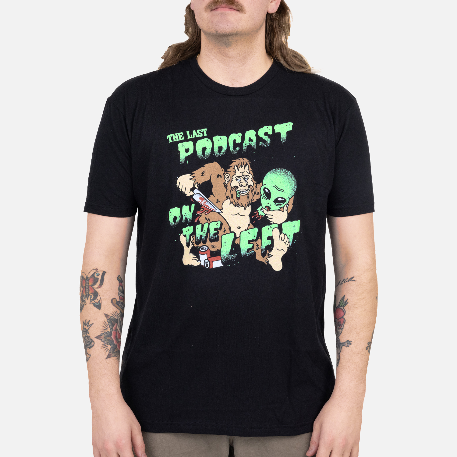 Black tee with graphic of bigfoot and alien head with text "THE LAST PODCAST ON THE LEFT" on male model