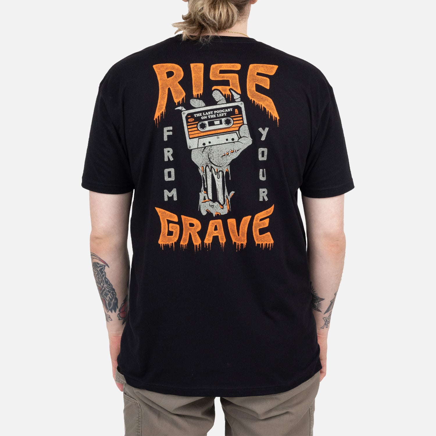 Rear view of black shirt on male model with graphic of skeleton hand holding cassette with text "THE LAST PODCAST ON THE LEFT" surrounded by text "RISE FROM YOUR GRAVE"