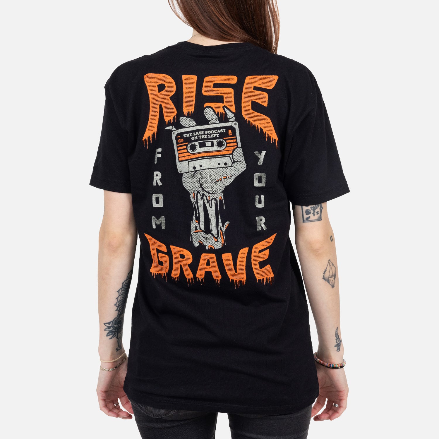 Rear view of black shirt on female model with graphic of skeleton hand holding cassette with text "THE LAST PODCAST ON THE LEFT" surrounded by text "RISE FROM YOUR GRAVE"