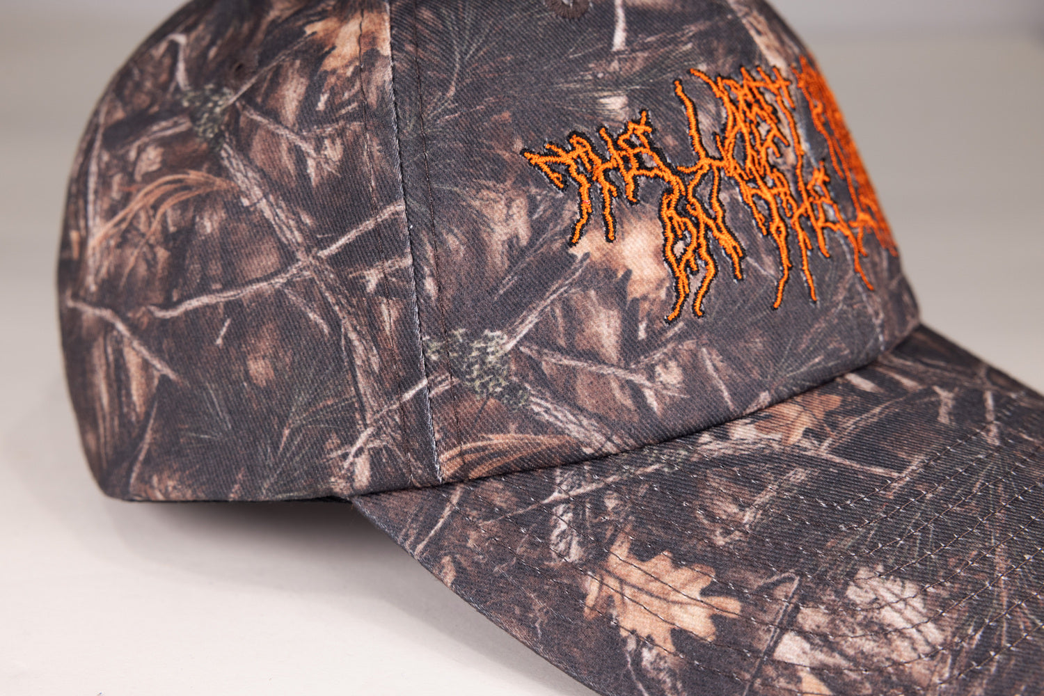 Closeup of Camo hat with orange death metal stylized text "THE LAST PODCAST ON THE LEFT"