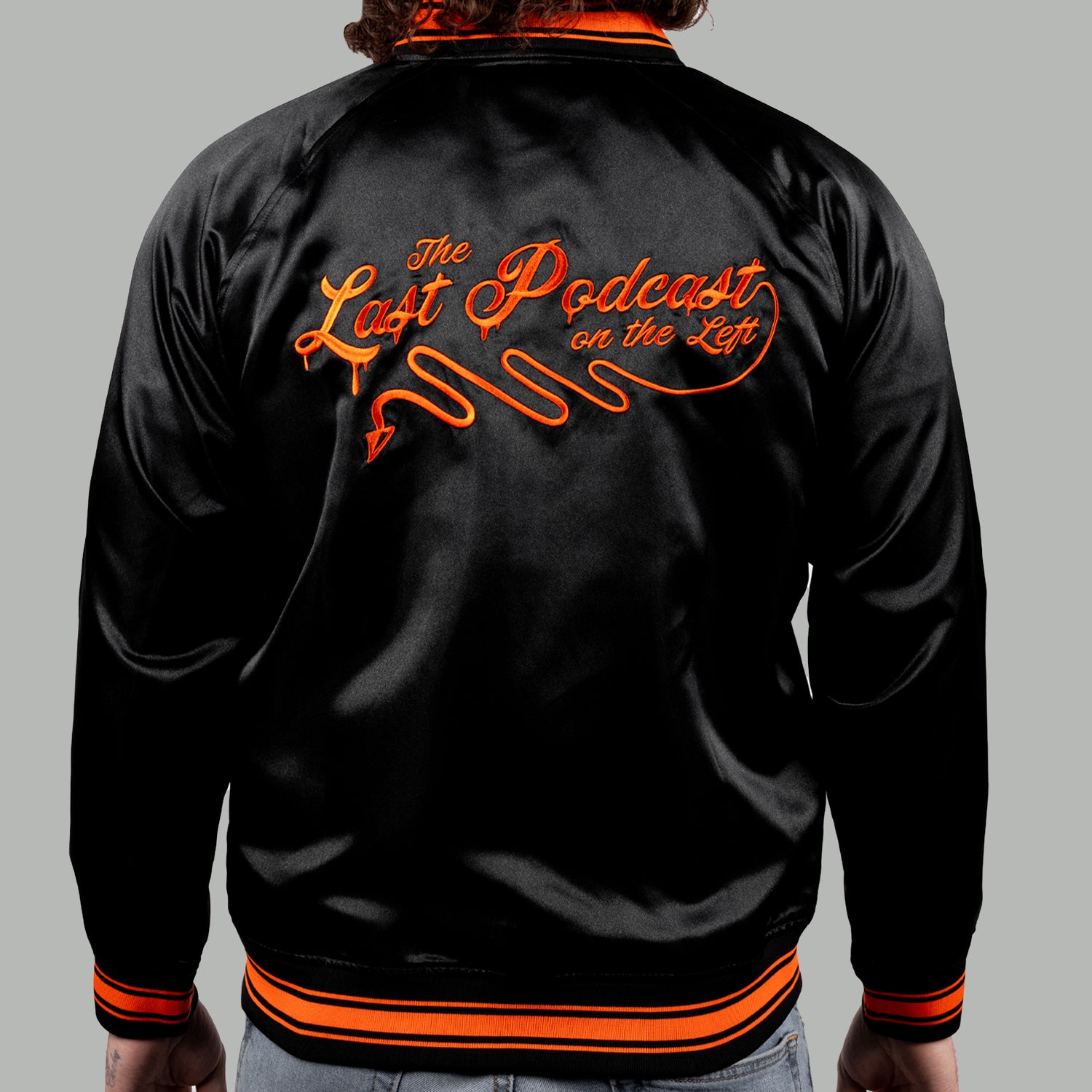 back of man in black satin jacket with orange text on back "The Last Podcast on the Left"
