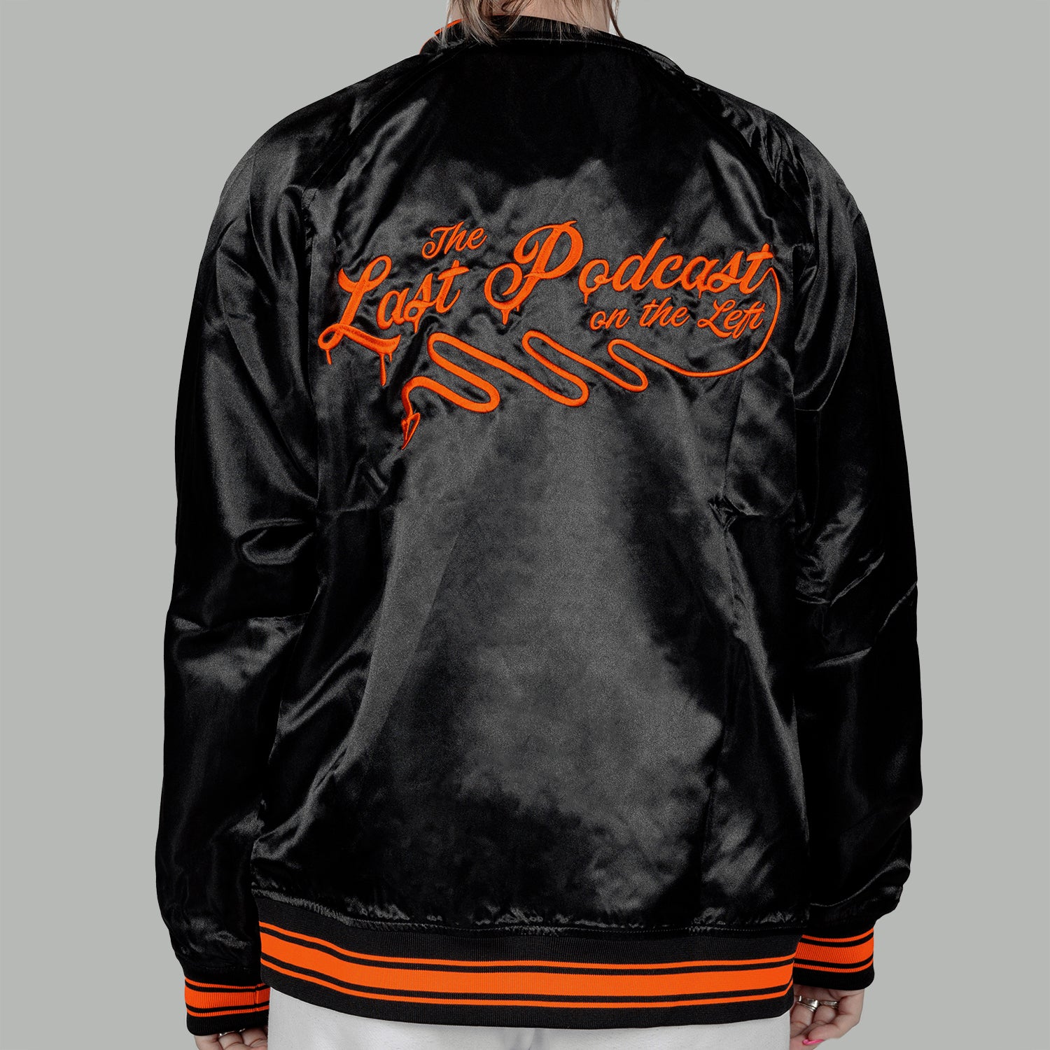 back of woman in black satin jacket with orange text on back "The Last Podcast on the Left"