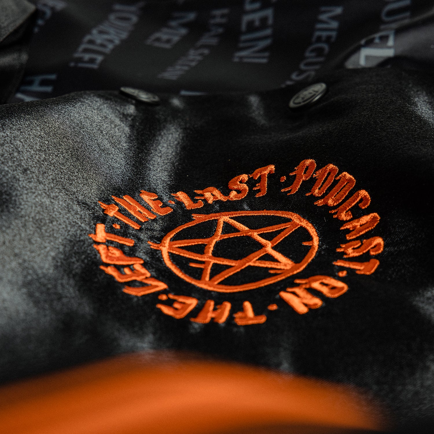 closeup of front left of black satin jacket showing orange pentagram with text "THE LAST PODCAST ON THE LEFT"