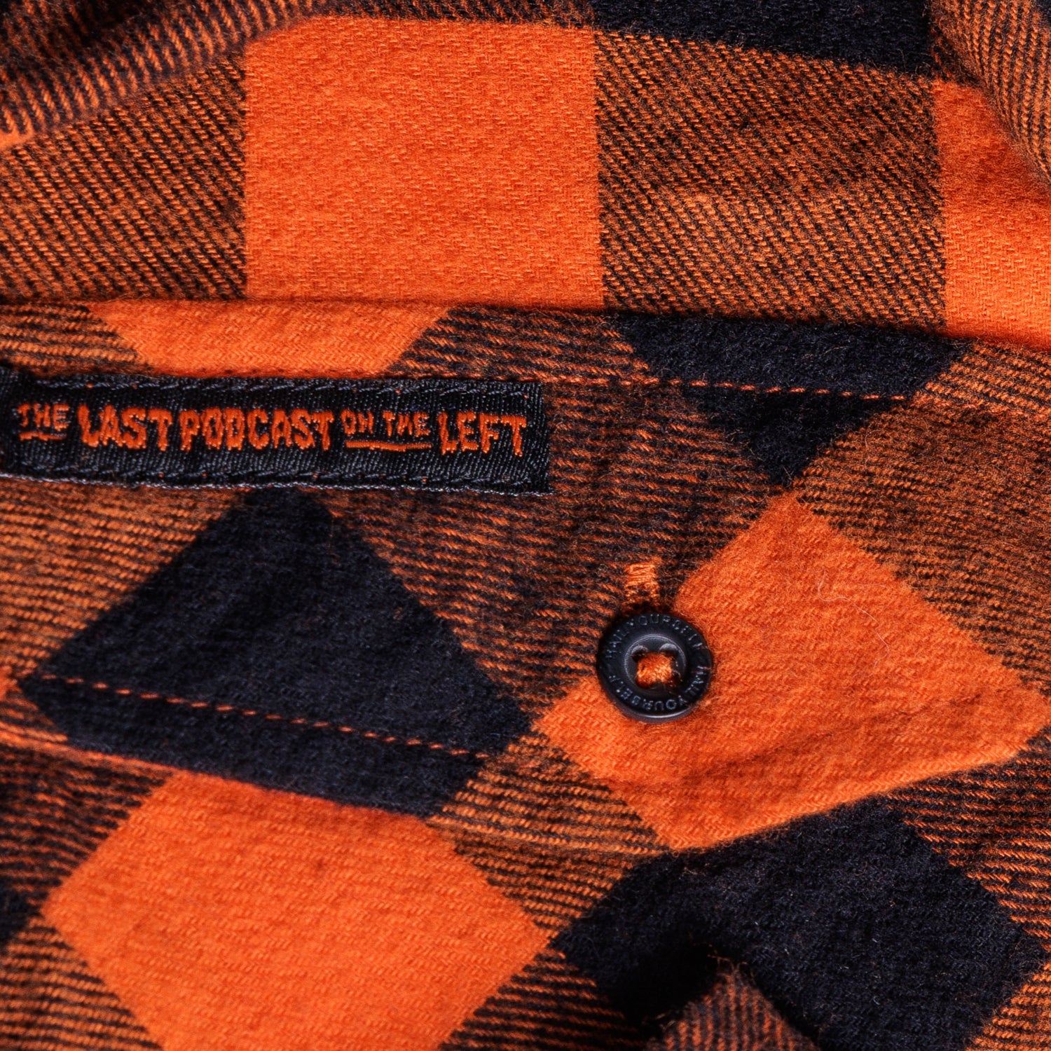 closeup of pocket of orange/black flannel buttondown with label with orange text "THE LAST PODCAST ON THE LEFT"