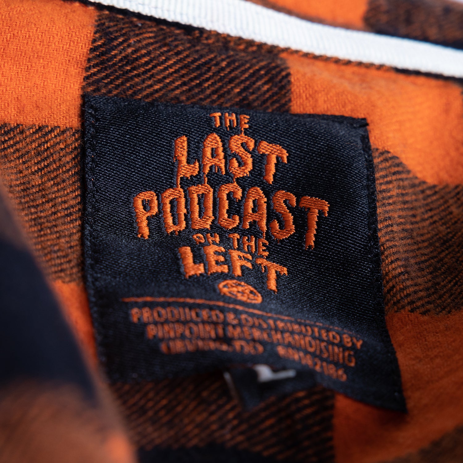 closeup of label on orange/black flannel buttondown black label with orange text "THE LAST PODCAST ON THE LEFT"