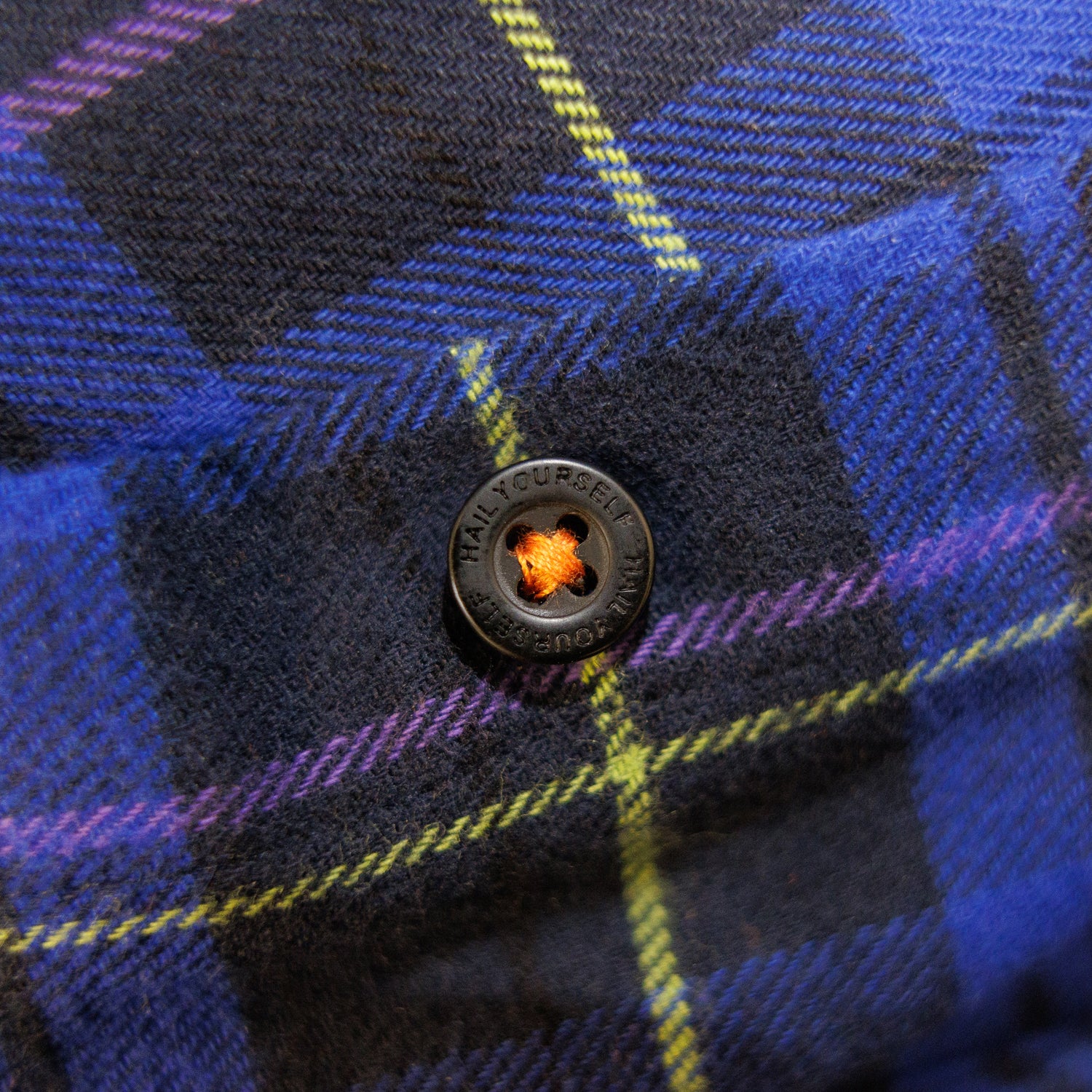closeup of button on blue plaid flannel buttondown, showing black button with orange thread