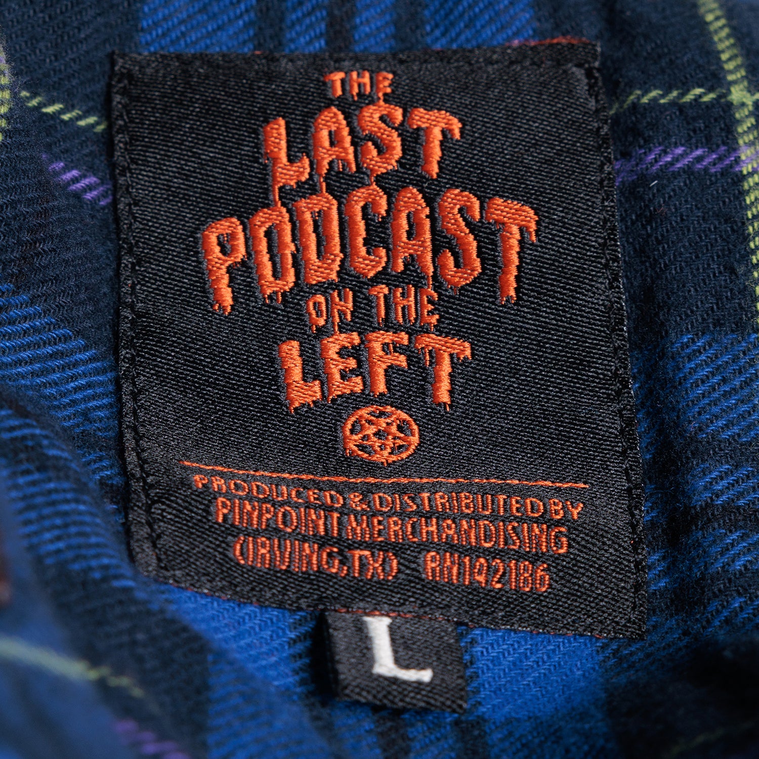 closeup of label on blue plaid flannel buttondown with orange text "THE LAST PODCAST ON THE LEFT"