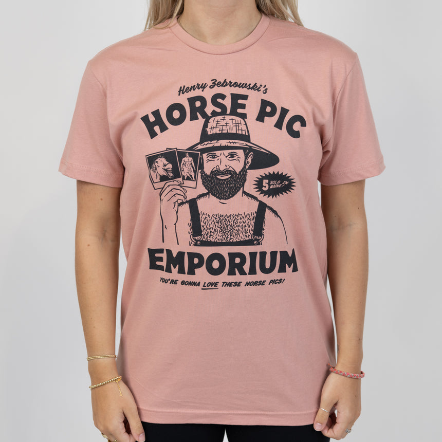 Man in desert pink shirt with black graphic of shirtless man in overalls and straw hat holding 2 polaroids of horses surrounded by text "Henry Zebrowski's HORSE PIC EMPORIUM YOU'RE GONNA LOVE THESE HORSE PICS! 5 SOLD IN MAINE!"